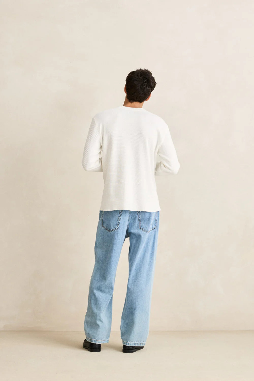 back of a man wearing a rhythm classic waffle knit white sweater and light blue jeans against a beige background