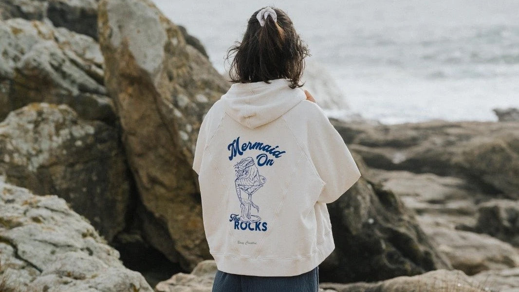 Person wearing an oversized Bauy hoodie with text and graphics, standing on a rocky beach.