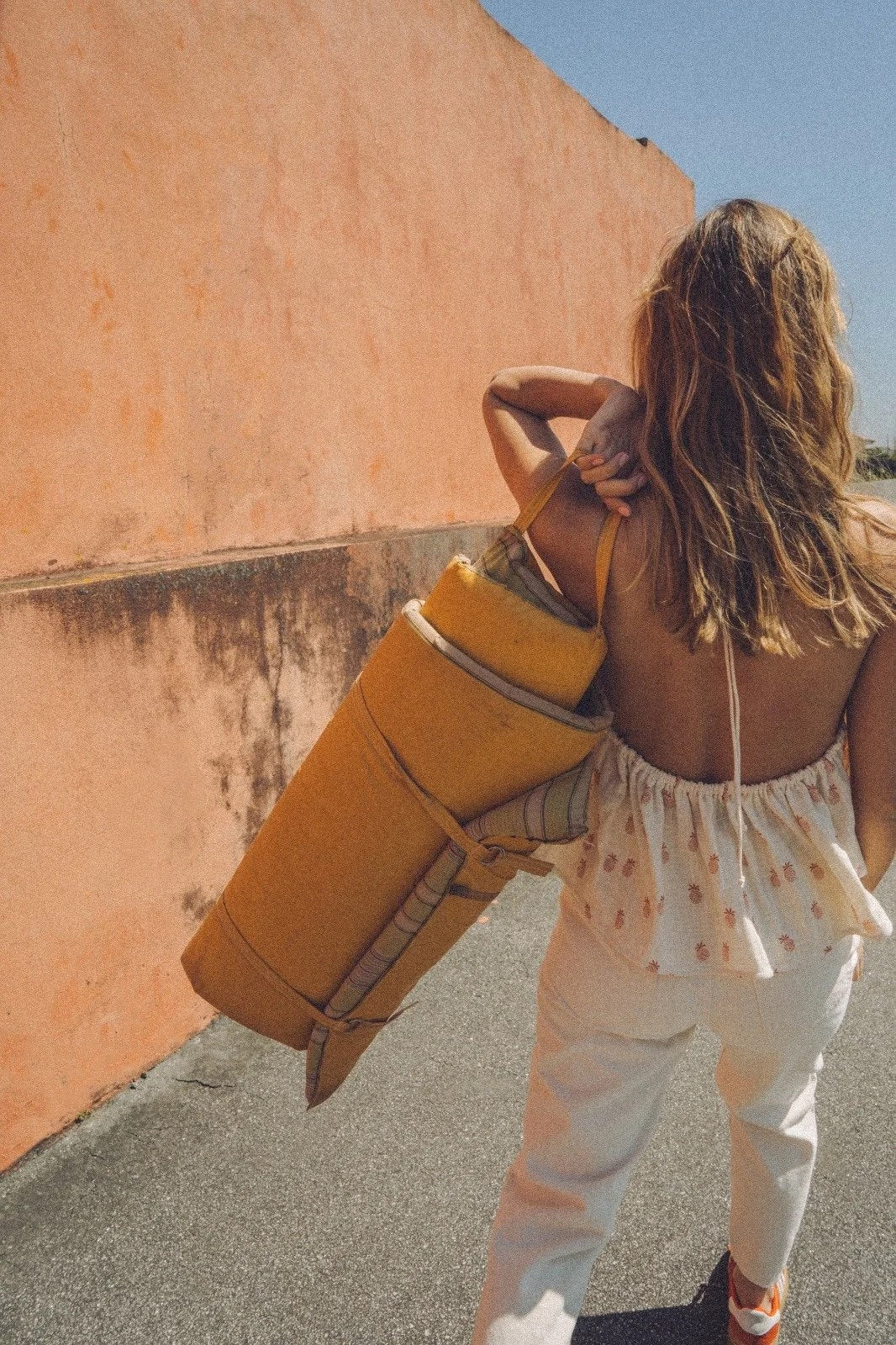 Woman with a yellow backpack walking along a street with an orange wall.