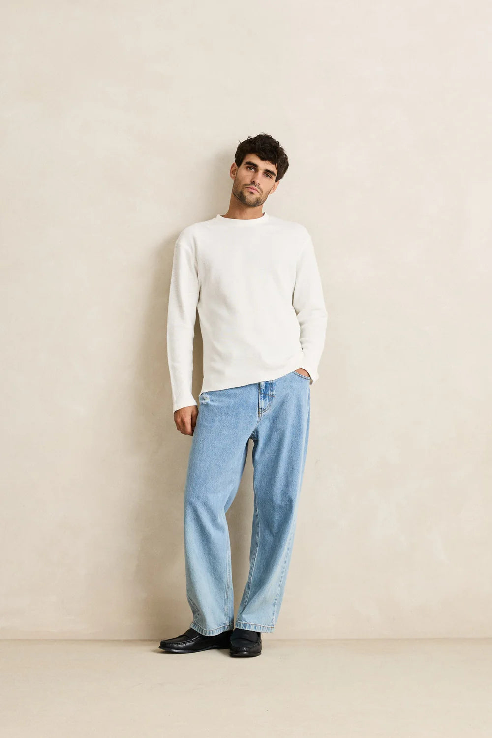 Man wearing a rhythm classic waffle knit white sweater and light blue jeans against a beige wall.