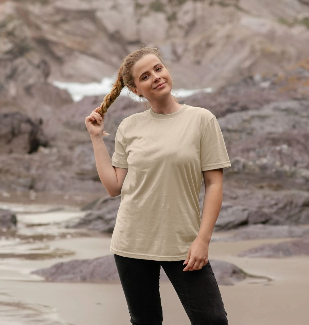 Woman standing on a rocky beach wearing a beige t-shirt and black pants.