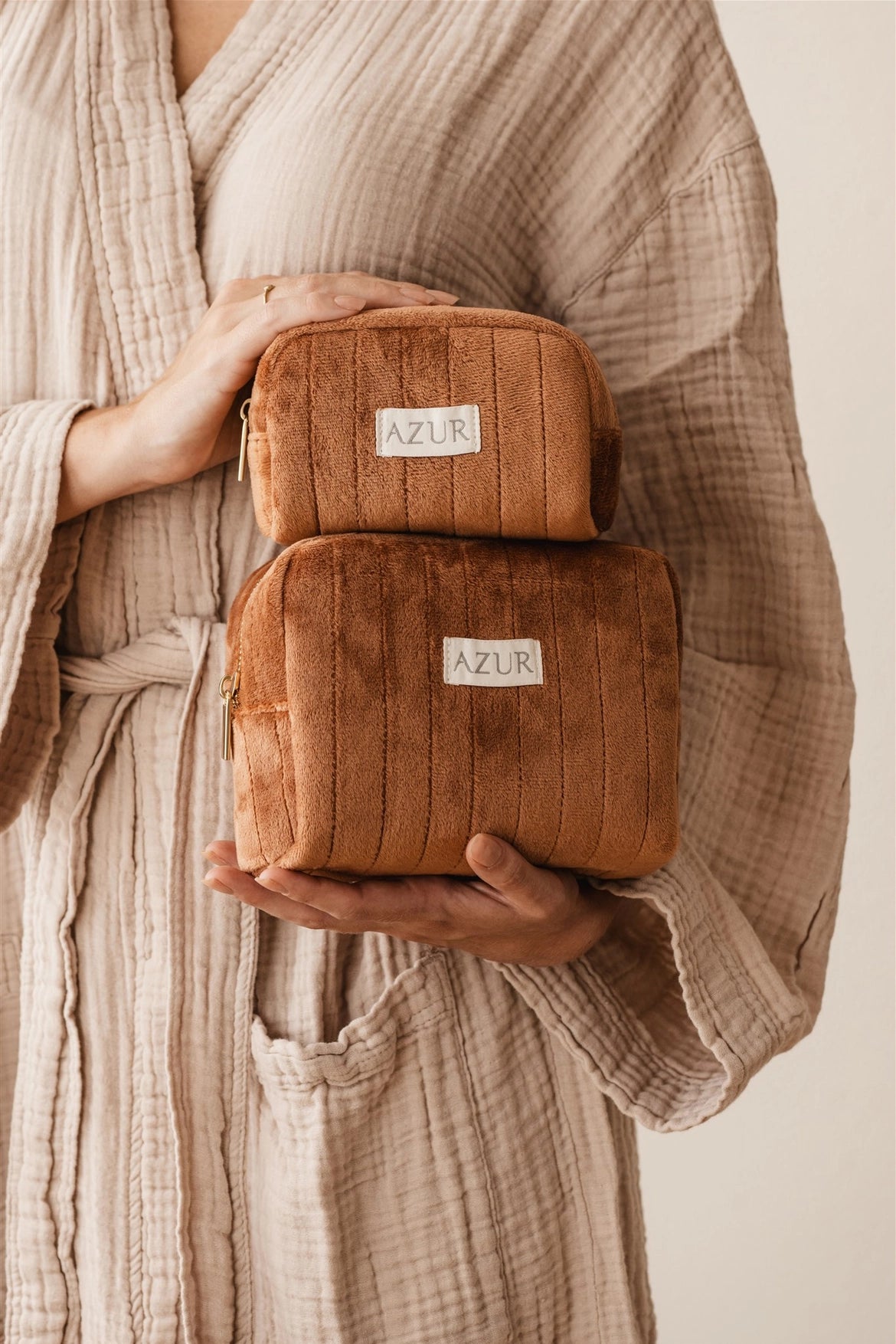 Person holding two brown toiletry bags with 'AZUR' label, wearing a beige robe.