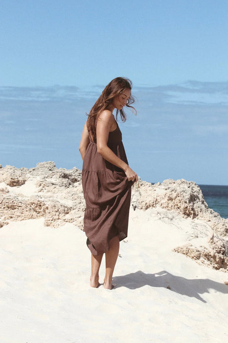 Woman in a rhythm chocolate tiered maxi dress standing on a rocky beach with clear blue sky.