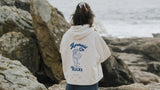 Person wearing an oversized Bauy hoodie with text and graphics, standing on a rocky beach.