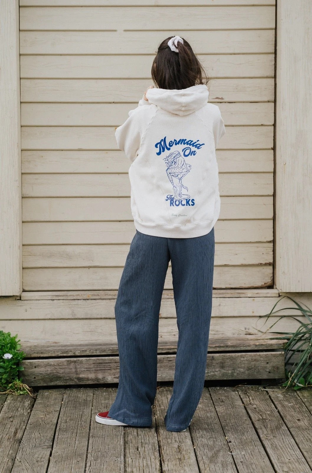 Person wearing a Bauy oversized white hoodie with a 'Mermaid On Rocks' design, standing in front of a wooden wall.