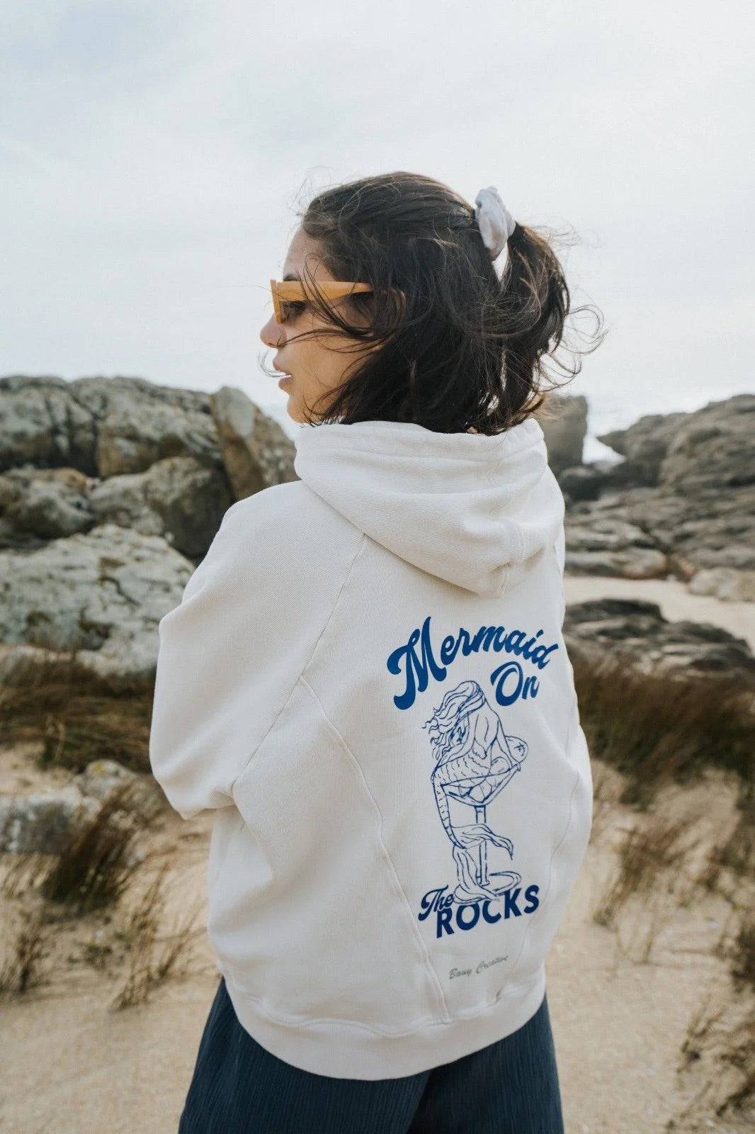 Person wearing a Bauy oversized hoodie with 'Mermaid on Rocks' design in a coastal setting