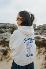 Person wearing a Bauy oversized hoodie with 'Mermaid on Rocks' design in a coastal setting