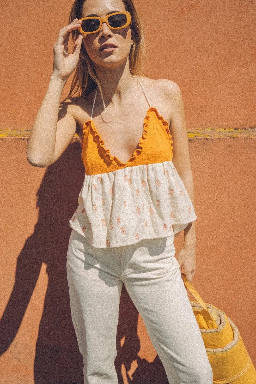 Woman wearing an orange Bauy pineapple top and white pants against a red wall