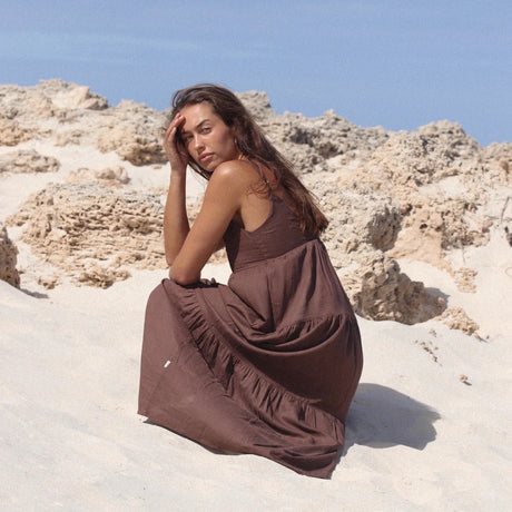 Woman in a brown dress sitting on a rocky beach with clear blue sky.