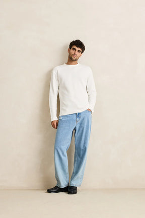 Man wearing a rhythm classic waffle knit white sweater and light blue jeans against a beige wall.